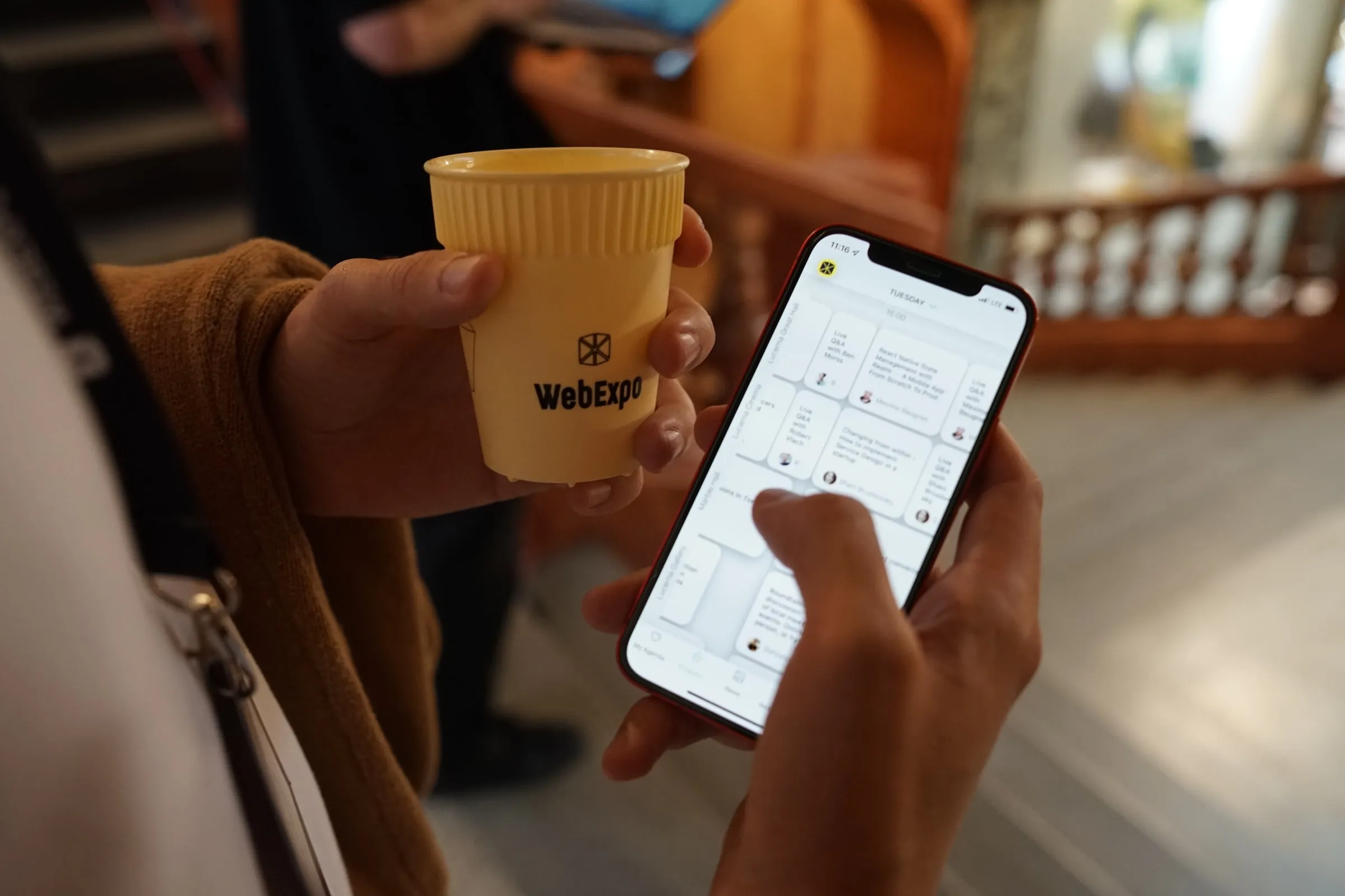 An attendee at WebExpo in Prague holding a coffee and browsing the conference agenda in the Eventee app on an iPhone